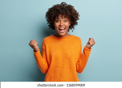 Joyful dark skinned model with crisp hair, clenches fists, feels overjoyed after winning game, wears orange jumper, poses over blue background, expresses good emotions. People and success concept