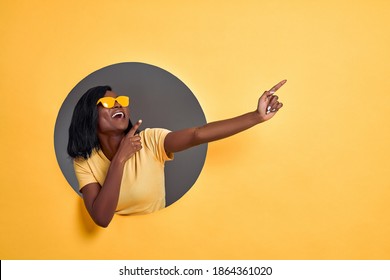 Joyful dark skinned woman points fingers aside , pleased by big sale prices, wears sunglasses and casual orange t shirt, models over yellow background, shows copy space for your advert.