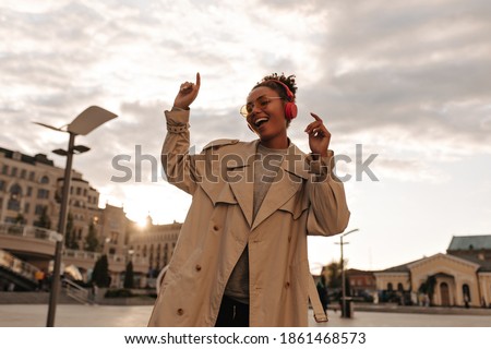 Joyful dark-skinned woman in oversized beige trench coat and eyeglasses smiles, listens to music and dances outdoors.
