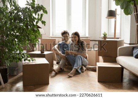 Joyful dating couple moving in apartment together, enjoying relocation, sitting on floor, hugging at heap of paper boxes, using online app, service on mobile phone, smiling, laughing