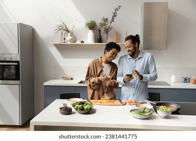 Joyful dating young African couple using online app on smartphones, cooking together in home kitchen, ordering food delivery on supermarket store internet website, consulting recipe on Internet - Powered by Shutterstock