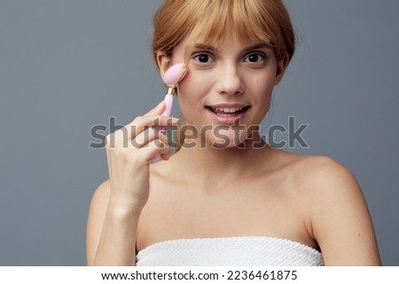 a joyful, delighted woman with perfect skin stands on a gray background and does a facial massage with a pink roller, smiling pleasantly. Close horizontal photo with empty space