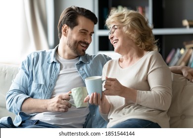 Joyful different generations bonding family relaxing on cozy sofa, drinking hot tea coffee, involved in funny conversation. Happy older 60s retired mother talking having fun with grown son at home.