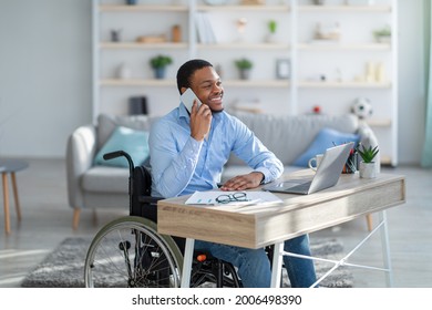 Joyful Disabled Black Guy In Wheelchair Having Phone Conversation While Working Online On Laptop From Home. Young Handicapped Businessman Talking With Client On Mobile Phone