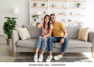 Joyful diverse couple watching TV program, sitting on couch at home, full length portrait. Cheery Arab man and his wife with remote control enjoying interesting movie in living room