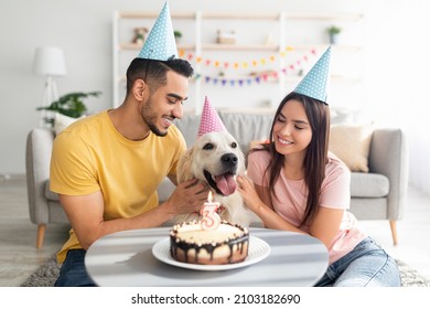 Joyful diverse spouses celebrating their dog's birthday with tasty cake, wearing festive hats at home. Adorable golden retriever having b-day party with owners. Holiday celebration concept - Powered by Shutterstock