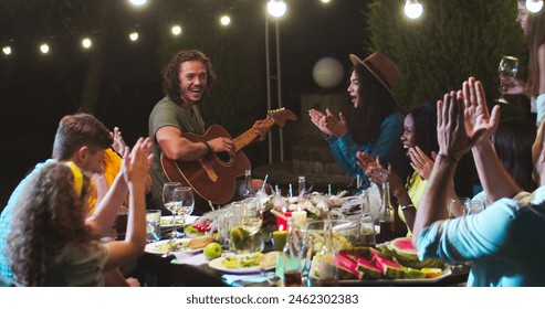 Joyful diverse young people having fun at party in garden at summer evening. Happy Caucasian male playing the guitar and singing outdoor. African American and Asian girls applaud. Leisure concept - Powered by Shutterstock