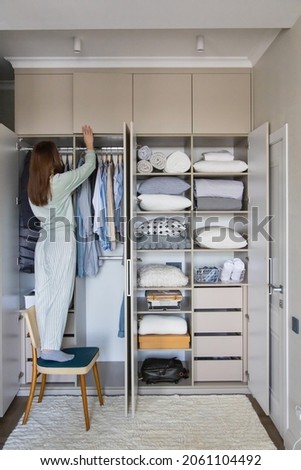 Joyful domestic female putting box on shelf in cupboard enjoying housework at wardrobe. Happy housewife organizing storage in closet at comfortable home. Smiling woman in pajamas arranging clothes