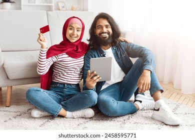 A joyful Eastern couple is casually seated on the floor, using digital tablet. The woman, wearing a red hijab and striped shirt, holds a credit card, suggesting a shopping or financial activity - Powered by Shutterstock