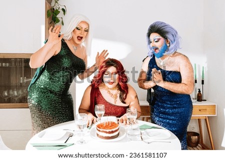 Joyful eccentric male drag queens in wigs and festive dresses with bright makeup screaming happily while standing near friend blowing candles on cake during birthday celebration