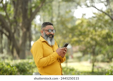 Joyful elder bearded man using mobile phone in garden. Cheerful bearded man stands on the pubic park and using by phone