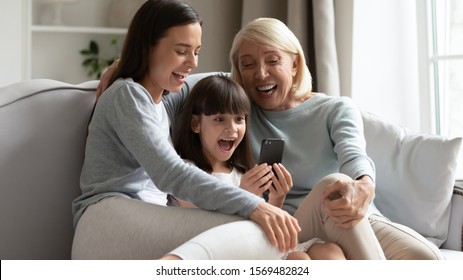 Joyful elderly 60s grandmother adult grown up daughter and granddaughter having fun on-line, small kid girl holding smart phone sitting between beautiful mom and grandma enjoy new funny apps concept - Powered by Shutterstock