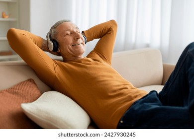 Joyful elderly grey-haired man reclining on couch in cozy living room, using wireless headphones, listening to music with closed eyes, grandfather relaxing at home, copy space - Powered by Shutterstock