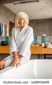 Joyful Elderly Lady In Bathrobe Sitting On The Edge Of Bathtub Stock Photo