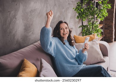 Joyful elderly woman celebrating with a smartphone while sitting on a cozy sofa in a modern home interior, wearing a blue sweater - Powered by Shutterstock