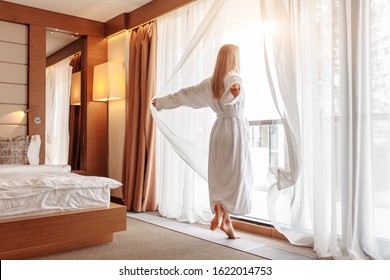 Joyful emotional young beautiful woman in white bathrobe happily opens curtains of her window in bedroom on sunny summer morning. The concept of starting a new day and fulfilling your plans - Powered by Shutterstock