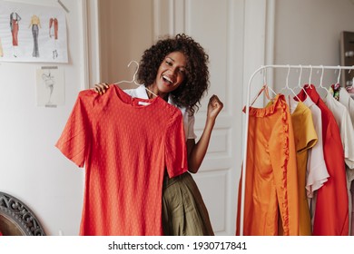 Joyful Energized Brunette Curly Dark-skinned Woman In Khaki Shorts Rejoices And Holds Hanger With Red Dress In Dressing Room.