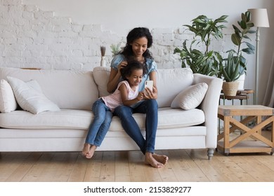 Joyful Engaged Black Mom Teaching Cute Preschool Daughter Kid To Use Online Learning App On Mobile Phone. Millennial Mother And Kid Shopping On Internet, Chatting, Talking On Video Call In Living Room