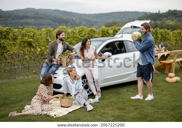 Joyful european friends on picnic near vineyards\
in countryside. Young men and women spend time together. Concept of\
winemaking and green tourism. Friendship. People with fresh\
watermelon and grapes