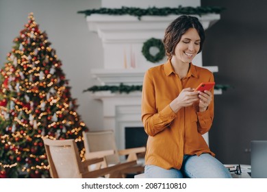 Joyful european woman using cellphone while sitting home office with Christmas tree on background, smiling hispanic female checking email or news feed of social network account during xmas holidays - Powered by Shutterstock