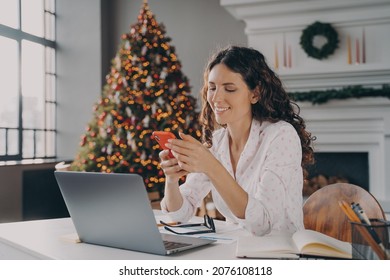 Joyful europian woman sitting at desk with laptop, reading xmas news or message on smartphone while working remotely in room with christmas decoration, happy female shopping online via mobile phone - Powered by Shutterstock