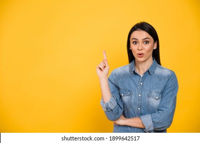 Joyful excited brunette young woman dressed in casual stylish blue shirt, points finger up, with surprised face, standing on isolated orange background, copy space, mock up