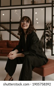 Joyful Excited Woman In Stylish Silk Pants And Black Jacket Sits On Leathered Brown Chair At Home. Brunette Lady In Suit And Eyeglasses Smiles In Living Room.