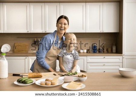 Joyful excited young mom and pretty little daughter girl preparing sandwiches in kitchen together, cutting ingredients, looking at camera, smiling, laughing, posing for cooking blog picture