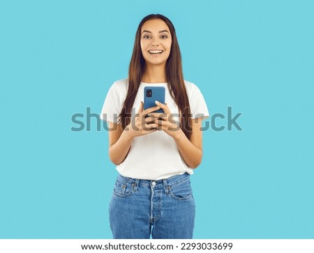Joyful excited young woman using mobile phone for online shopping, chatting, watching media or playing games. Girl with smiling shocked expression holding smartphone isolated on light blue background.
