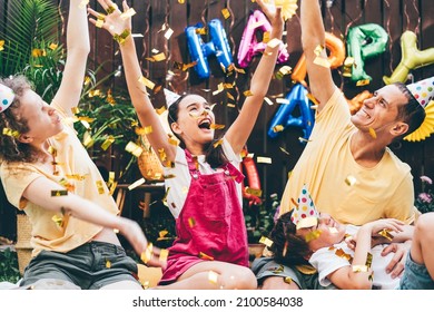Joyful family with children throws air balloons and golden confetti sitting on plaid at birthday celebration in decorated cottage yard in summer