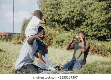 A joyful family enjoys a sunny day in the park, capturing memories with a camera. A child sits on a person's shoulders, surrounded by greenery and joy. - Powered by Shutterstock