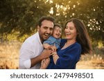 A joyful family moment with a father, mother in a blue dress, and their one-year-old son in blue clothes, hugging and smiling in a grassy natural setting with a tree in the background