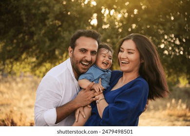 A joyful family moment with a father, mother in a blue dress, and their one-year-old son in blue clothes, hugging and smiling in a grassy natural setting with a tree in the background - Powered by Shutterstock