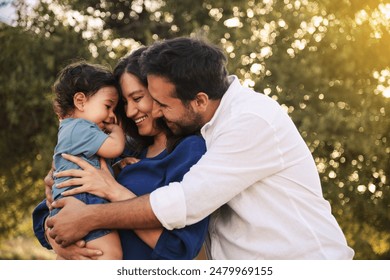 A joyful family moment with a father, mother in a blue dress, and their one-year-old son in blue clothes, hugging and smiling in a grassy natural setting with a tree in the background - Powered by Shutterstock