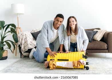 Joyful Family Multiracial Young Couple, Hispanic Guy And Caucasian Girl, Sit On The Floor At Home In The Living Room Near A Large Yellow Suitcase, Putting Clothes In A Suitcase For Vacation, Smiling