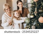 A joyful family scene of three generations decorating a Christmas tree together, capturing warmth, love, and festive spirit. Smiling women and child share a magical holiday bonding moment at home.