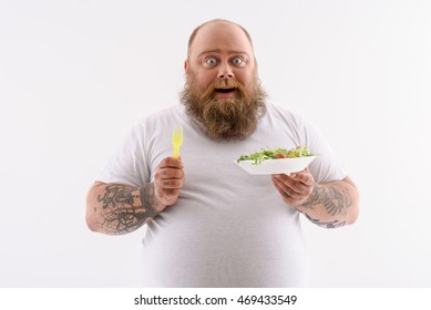 Joyful fat man is eating salad and smiling. He is standing and looking at camera with amazement. Isolated