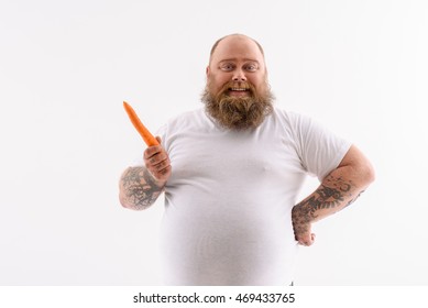 Joyful fat man is holding carrot and smiling. He is standing and looking at camera happily. Isolated