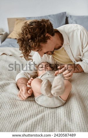 joyful father with curly hair and beard looking at his infant baby son on bed, precious moments