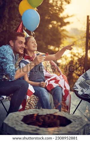 A joyful father and his daughter, both wearing  birthday hats,sitting beside a bonfire. Their faces are filled with excitement as they eagerly point towards someone who has just arrived at the party. 
