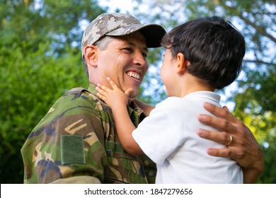 Joyful Father Holding Little Son In Arms, Hugging Boy Outdoors After Returning From Military Mission Trip. Low Angle. Family Reunion Or Returning Home Concept