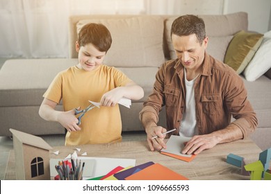 Joyful Father And Son Are Making Origami Together At Home. Boy Is Cutting Paper By Scissors While Man Is Drawing Shape