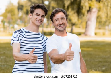 joyful father and teenager son in t-shirts showing thumbs up in park