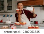 Joyful female baker using smart phone while making selfie with freshly made muffin on the kitchen