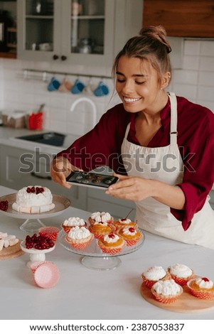 Joyful female baker using smart phone while photographing various desserts laying on the kitchen table