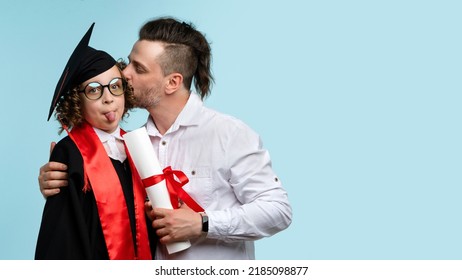 Joyful Female Child In Graduation Cap And Ceremony Robe Holds Certificate Shows Tongue Has Fun On Light Blue Studio Background. Dad Congratulates Prodigy Daughter On Graduating Elementary School