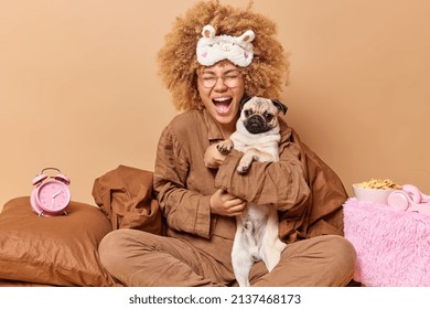 Joyful female pet owner poses in comfortable bed together with pug dog play and prepare for sleep pose in bedroom with alarmclock pillow blanket and bowl of cornflakes around. Domestic animals concept