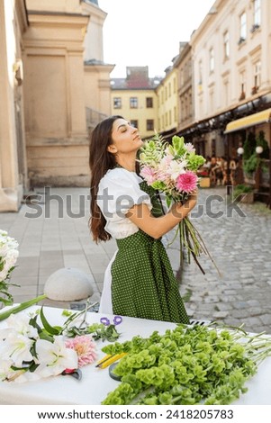 Joyful florist in green polka dot apron holds freshly made bouquet, savoring scent of her beautiful floral arrangement.