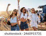 Joyful friends dance at sunset on sandy shore during summer music fest. Girls enjoy live band, seaside fun, wearing stylish outfits. Friendship, entertainment, youth culture at vibrant beach event.