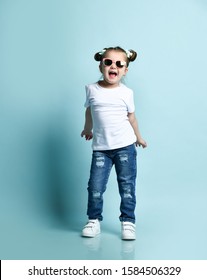 Joyful frolic smiling little baby kid girl with funny buns with bows and in sunglasses, t-shirt and blue jeans is dancing, playing, singing, shaking hips